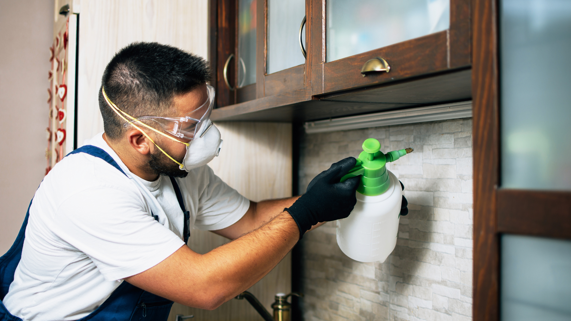 Pest control technician wearing protective mask and gloves spraying pesticide under kitchen cabinets in a residential property.